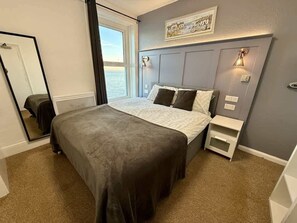 A bedroom with a bed, bedside tables, a mirror, and a view of the sea. - Fishermans Watch Apt. Oceana (East Riding of Yorkshire)