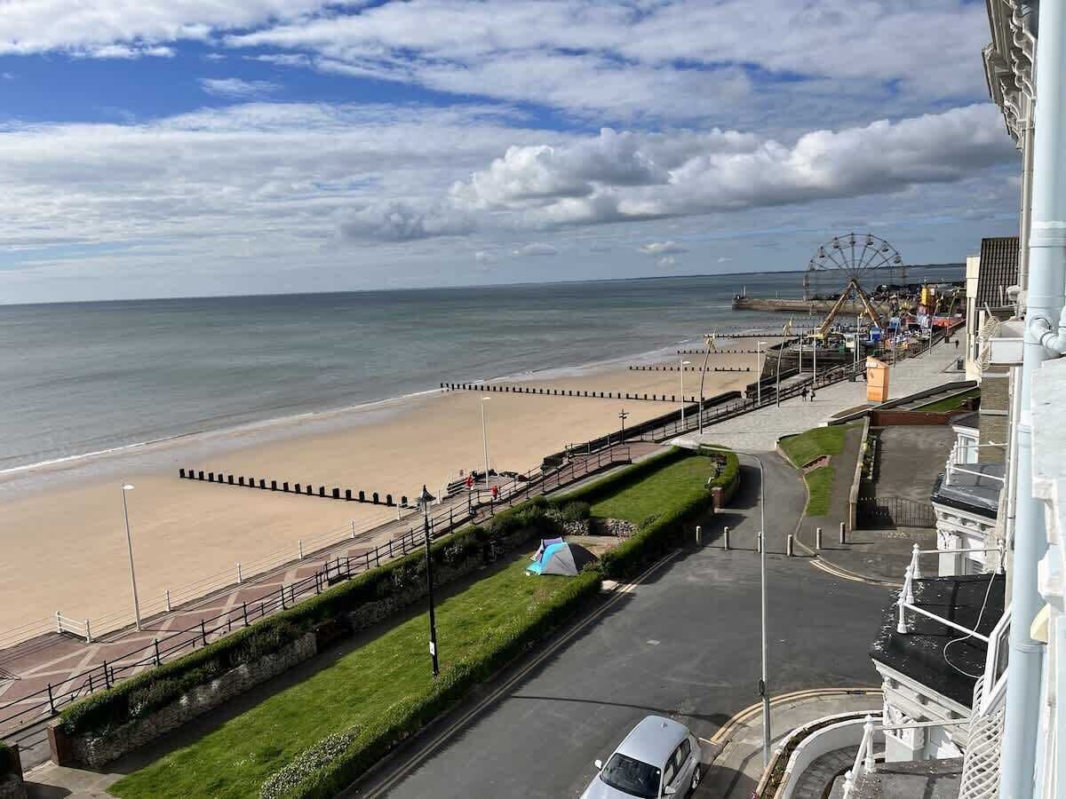 A coastal promenade with a beach, a fenced walking path, and a Ferris wheel in the distance.