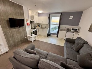 A modern living room with a grey sofa, a flat-screen TV mounted on the wall, and a kitchen area with white cabinets and a window. - Fishermans Watch Apt. Oceana (East Riding of Yorkshire)