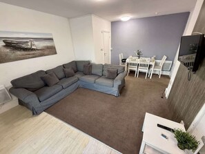 Ein Wohnzimmer mit einem grauen Ecksofa, einem kleinen weißen Tisch mit Pflanze und einem an der Wand montierten Fernseher.