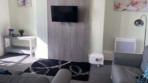 A living room with a flat-screen TV mounted on a wooden panel, a grey sofa, a white side table with a plant, and a black rug with a white pattern. - Promenade Apt. Oceana (East Riding of Yorkshire)