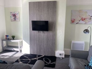 A living room with a flat-screen TV mounted on a wooden panel, a grey sofa, a white side table with a plant, and a black rug with a white pattern. - Promenade Apt. Oceana (East Riding of Yorkshire)