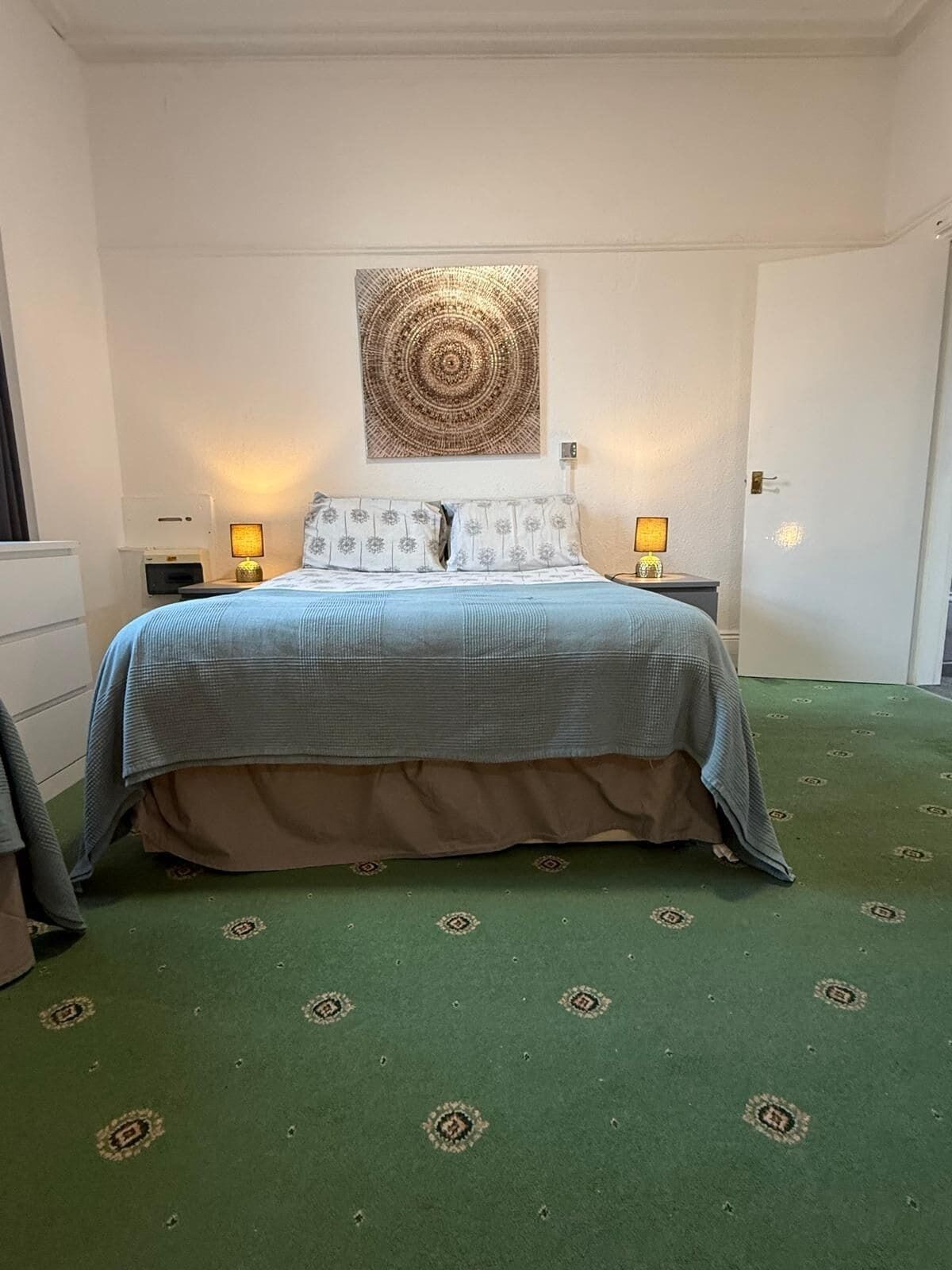 A hotel room with a bed, two bedside lamps, a wall-mounted artwork, and a patterned carpet.