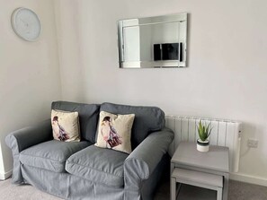 A grey sofa with two decorative pillows, a small bedside table with a potted plant, and a wall-mounted mirror. - Balcony sea view apt for couples (East Riding of Yorkshire)