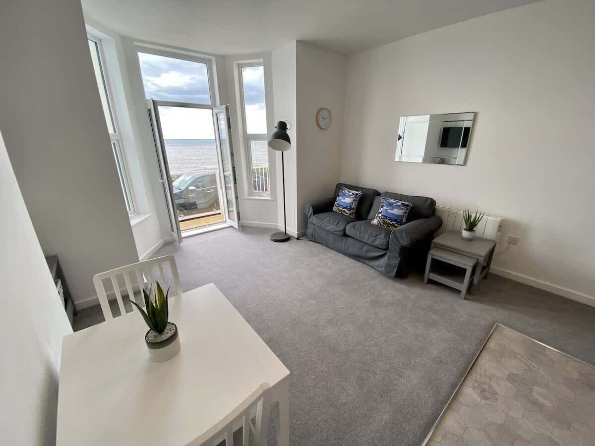 A modern living room with a grey sofa, a white table, and a view through large windows.