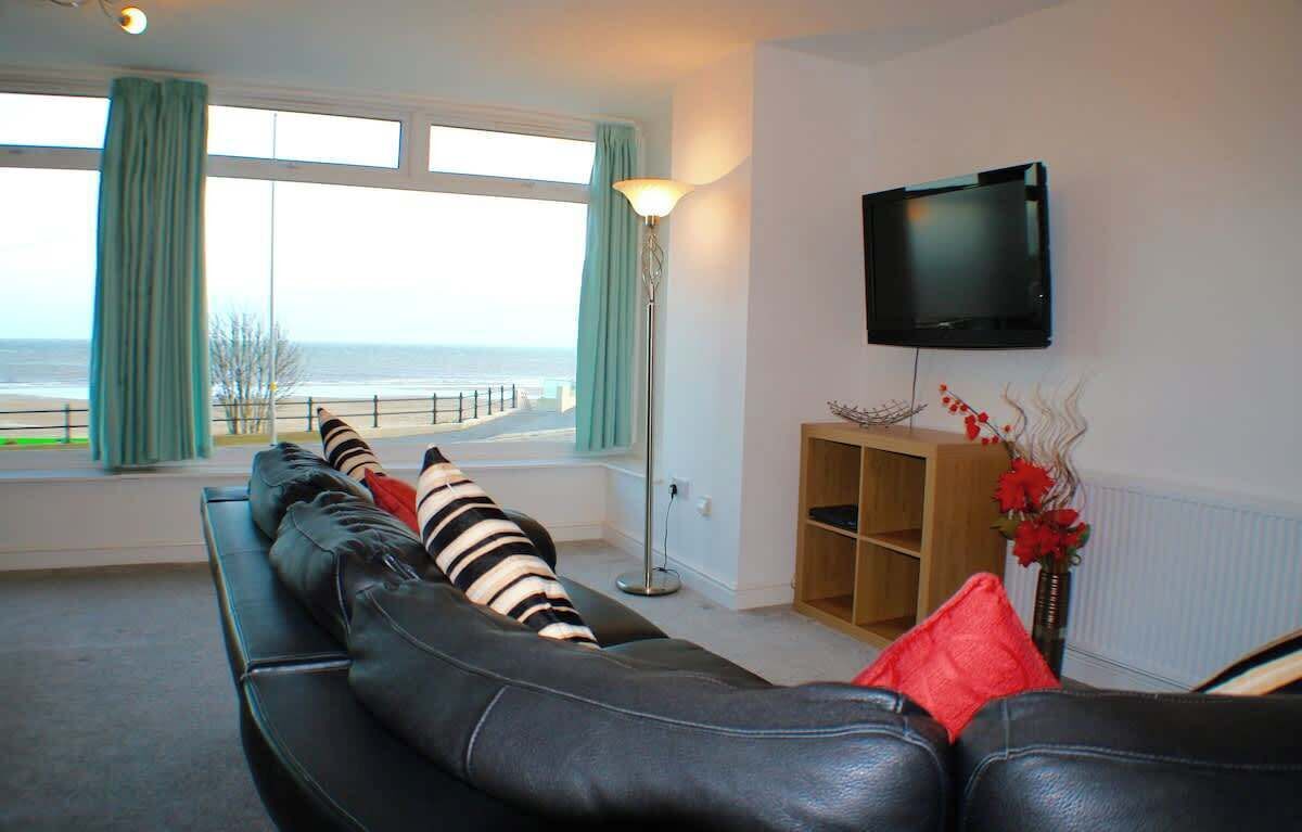 A living room with a sofa, a TV mounted on the wall, a floor lamp, and a view of the sea through large windows.