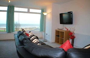A living room with a sofa, a TV mounted on the wall, a floor lamp, and a view of the sea through large windows. - 2 Bedroom Sea View Apartment with Balcony (Bridlington)