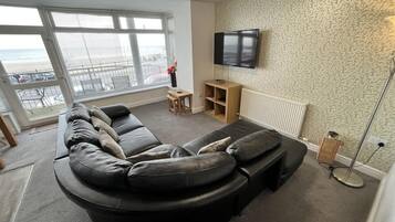 A living room with a curved black leather sofa, a flat-screen TV mounted on the wall, and a large window offering a view of the sea.