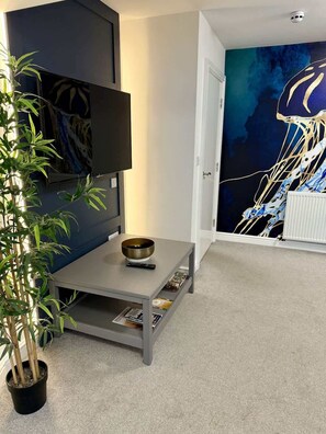A modern living room with a flat-screen TV, a small table with a bowl and remote, a potted plant, and a large abstract painting on the wall. - 2 bedroom apartment, sea views, balcony & parking (Bridlington)