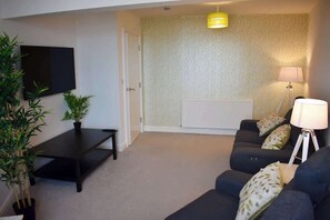 A living room with a sofa, a coffee table, a TV, a lamp, and two potted plants. - 2 bedroom apartment, sea views, balcony & parking (Bridlington)