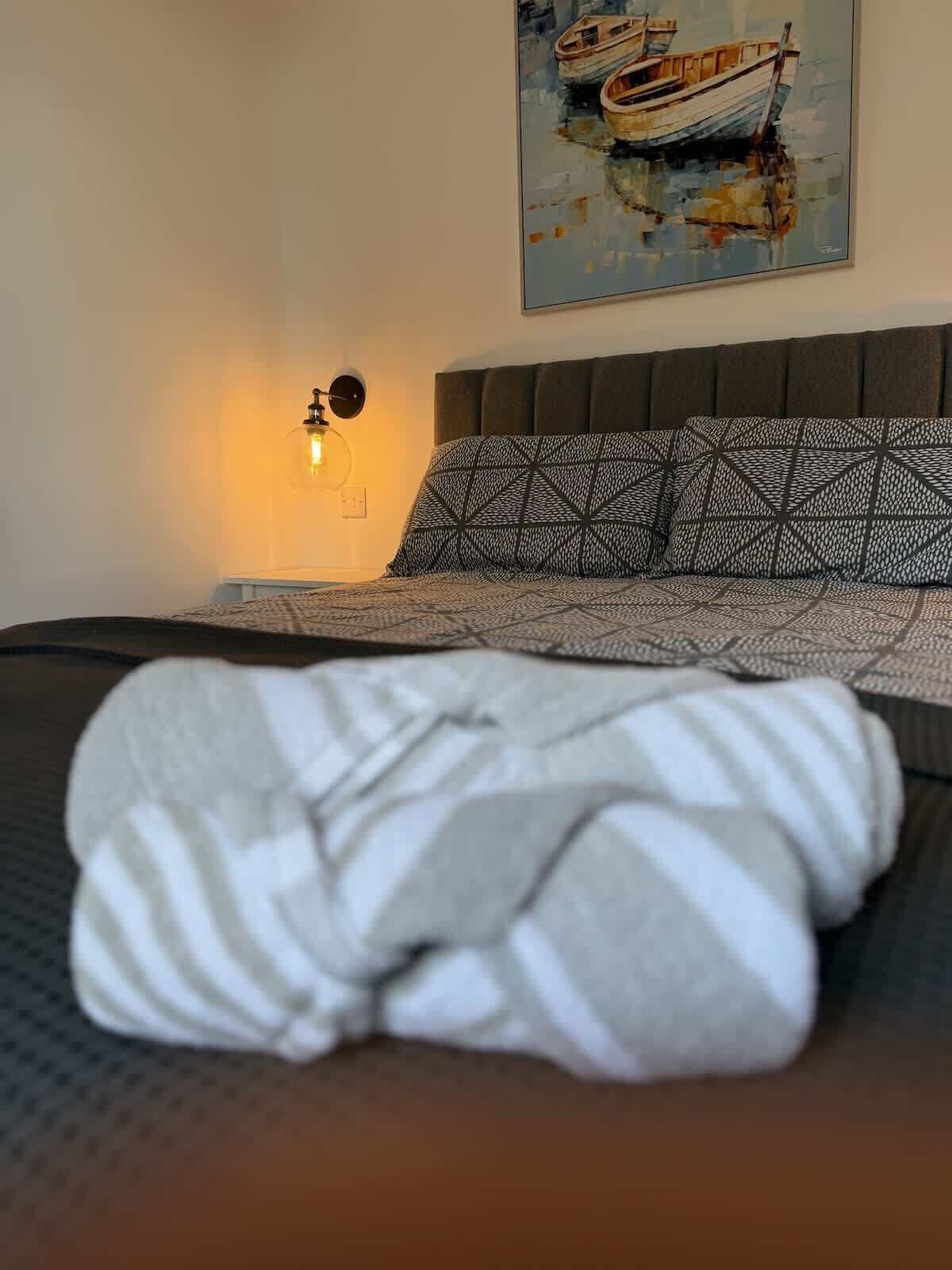 En pænt redt seng med pude med et mønster og et tæppe, en vægmonteret lampe og et maleri af en båd på væggen.