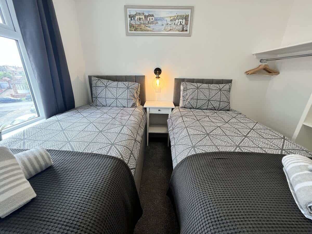 A room with two beds, a nightstand, a lamp, and a view of the outside.