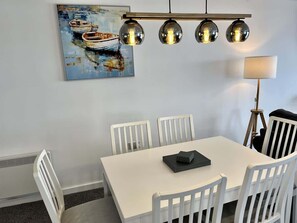 Et spiseområde med et hvidt bord, fire stole, et maleri af både og en moderne pendellampe.