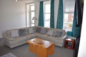 A living room with a beige sectional sofa, a wooden coffee table, and teal curtains. - Luxury 3 bedroom, sea view apartment with balcony (East Riding of Yorkshire)