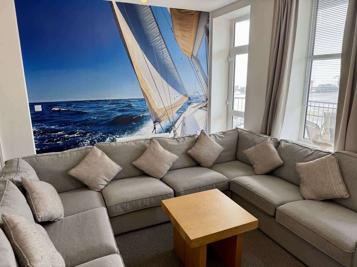 Un salon moderne avec un grand canapé d’angle, une table basse en bois et une fresque murale représentant un voilier.