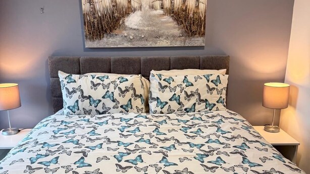 A bedroom with a bed, two nightstands, a wall-mounted painting, and two bedside lamps. - Ocean Wave Apt. Oceana (East Riding of Yorkshire)