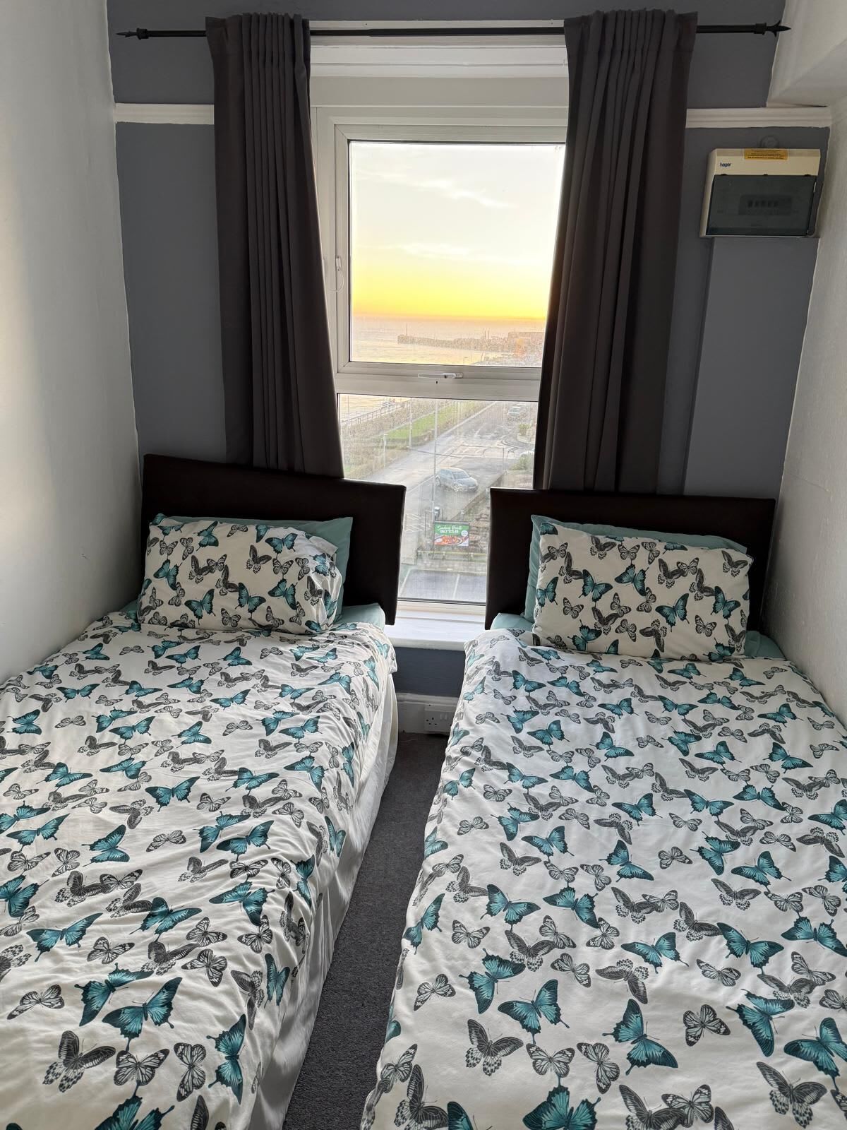 Habitación con dos camas, cada una con cubrecama estampado con mariposas, una ventana con cortinas y vistas a un paisaje urbano.