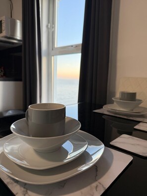 A kitchen with a view of the sea through a window, a microwave, and a set of white plates and cups. - Ocean Wave Apt. Oceana (East Riding of Yorkshire)