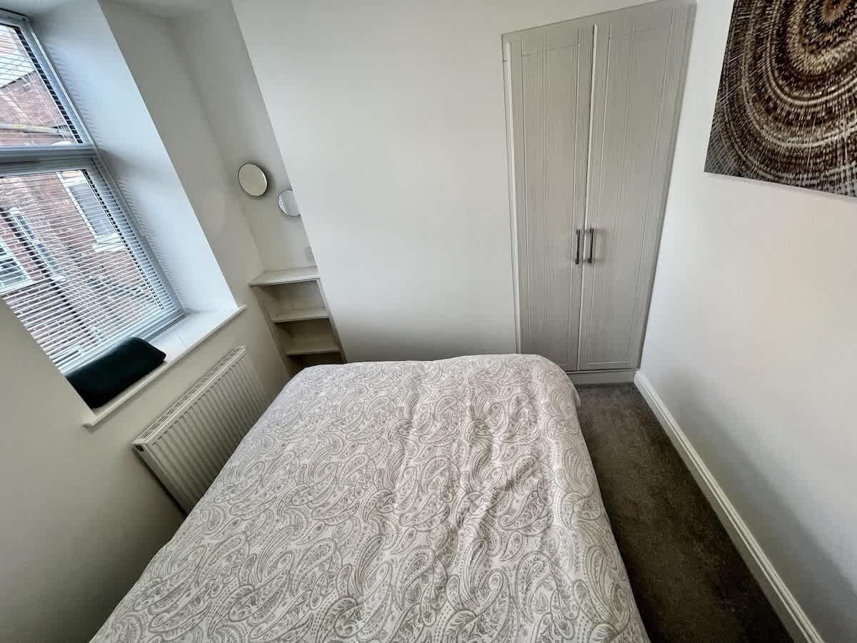 Een slaapkamer met een bed, een raam, een radiator, een kledingkast en een wandlamp.