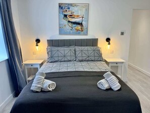 A neatly made bed with towels, a painting of boats on the wall, and two bedside lamps. - Seaside Escapes Apt. 2 (East Riding of Yorkshire)