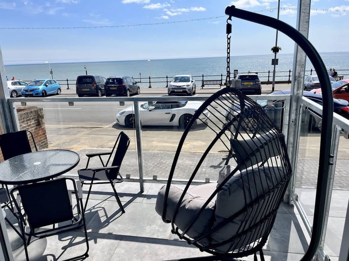 A balcony with a hanging chair, a table, and chairs, overlooking a parking area and the sea.