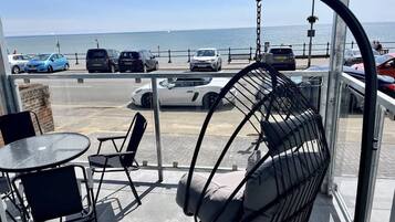 A balcony with a hanging chair, a table, and chairs, overlooking a parking area and the sea.