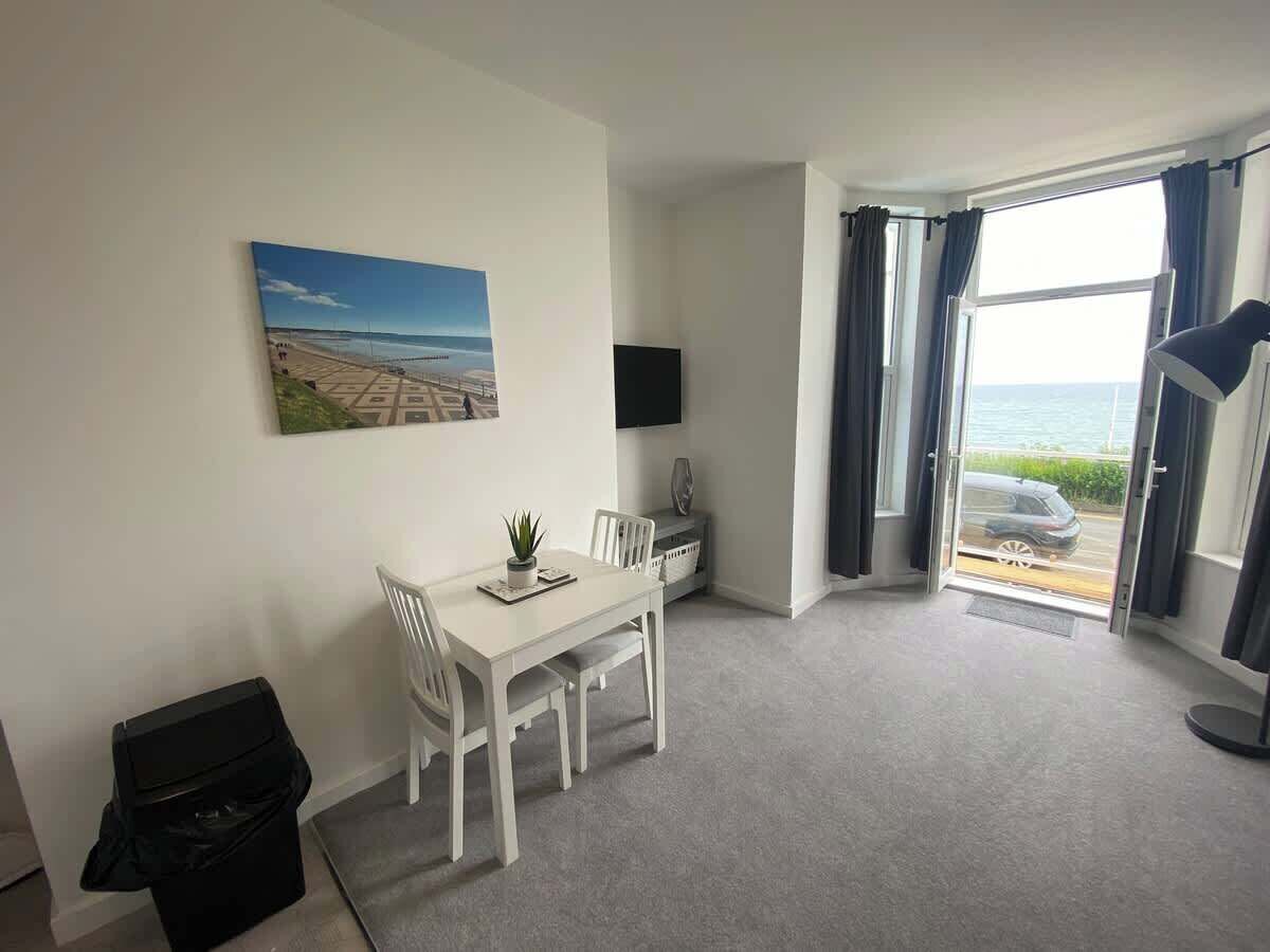 A room with a dining table, chairs, a TV, a window with curtains, and a view of the sea.