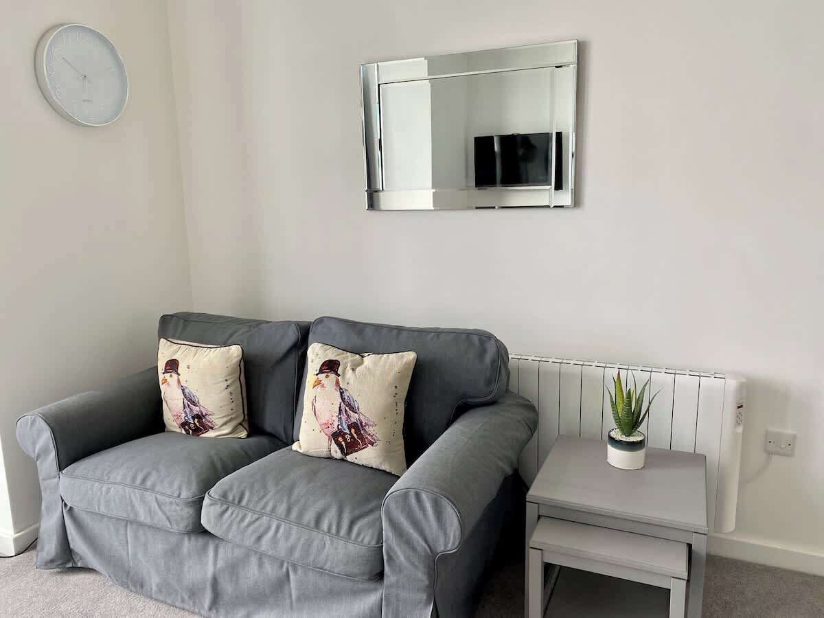 A grey sofa with two decorative pillows, a small bedside table with a potted plant, and a wall-mounted mirror.