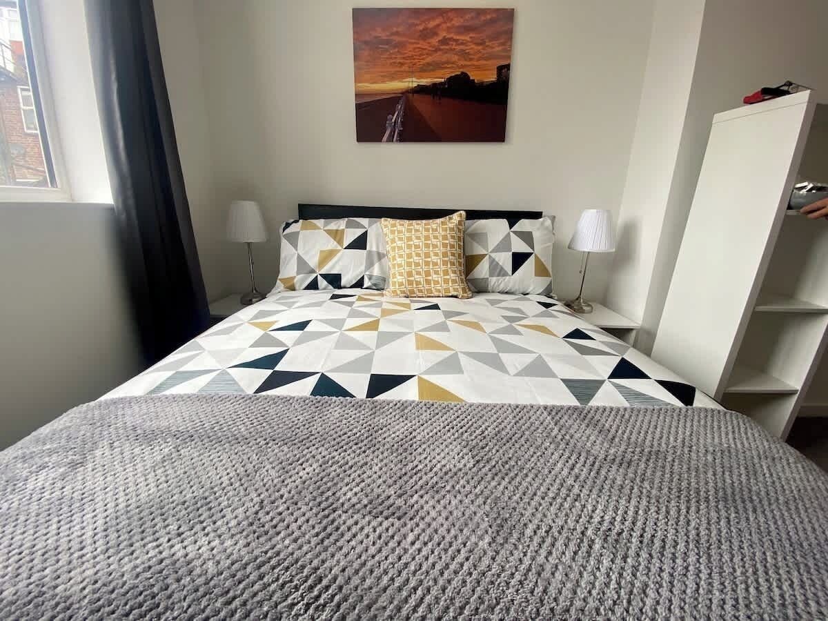 Ein Bett mit einem geometrisch gemusterten Überwurf, zwei Nachttischlampen und ein Sonnenuntergangsgemälde an der Wand.
