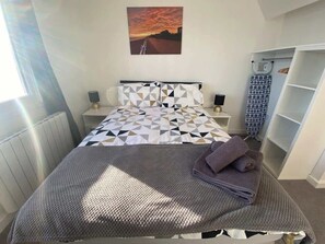 Ein Schlafzimmer mit Bett, Nachttisch, Lampe, einem Bild, einem Fenster und einem Kleiderschrank.