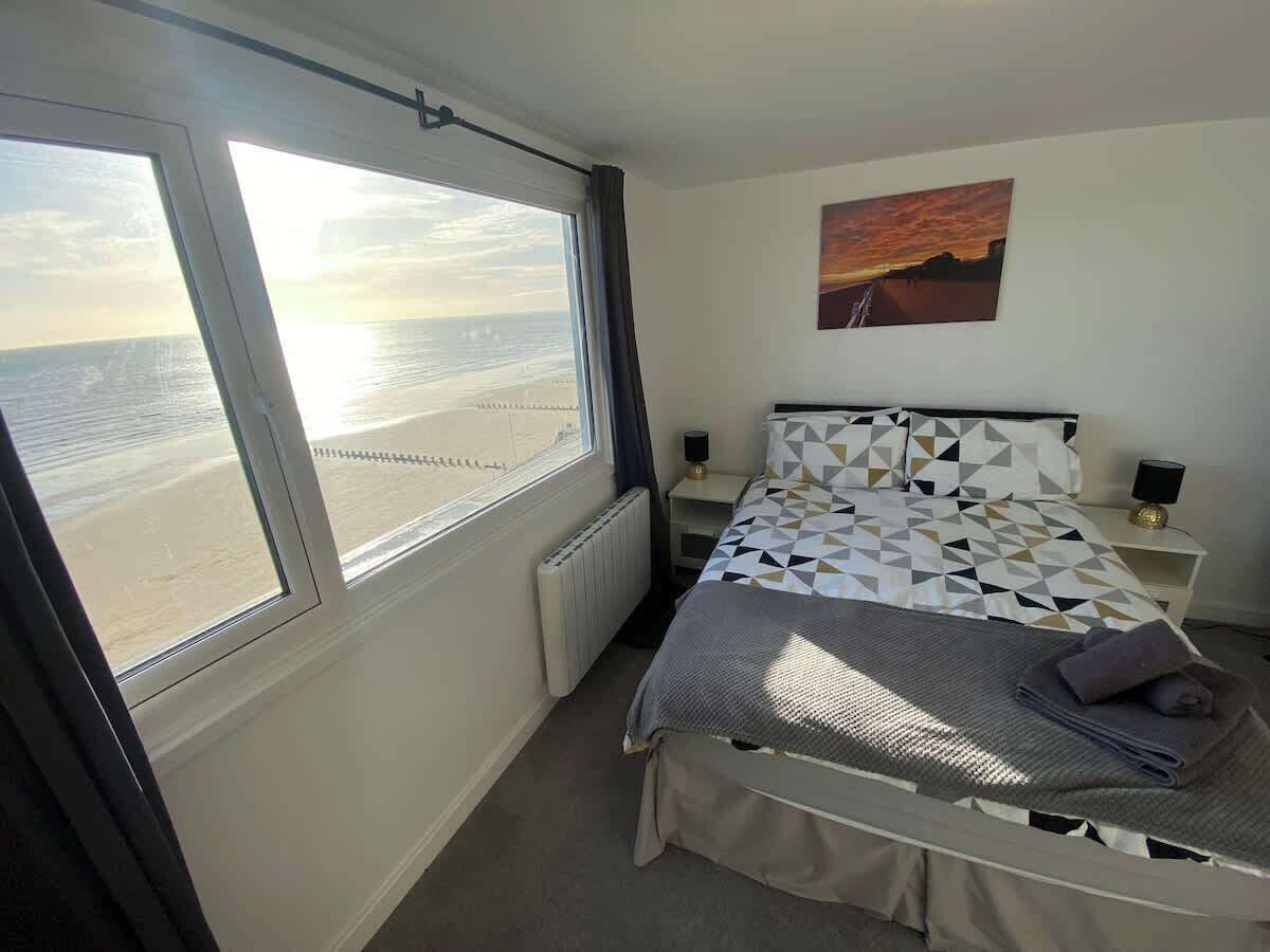 Un dormitorio con un ventanal grande, una cama con colcha estampada, una mesita de noche con lámpara y un cuadro en la pared.