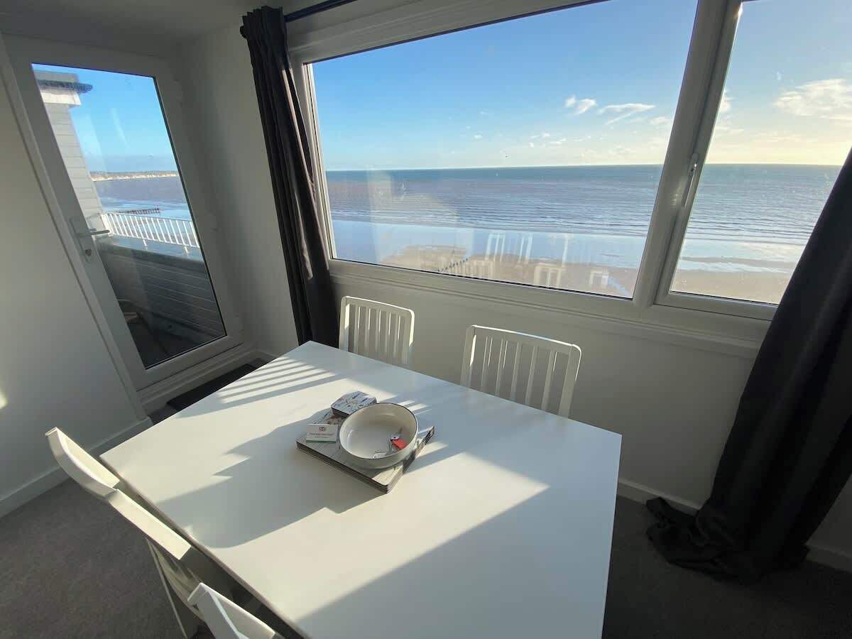 A room with a view of the ocean, a white table with a bowl and a book, and a window with curtains.