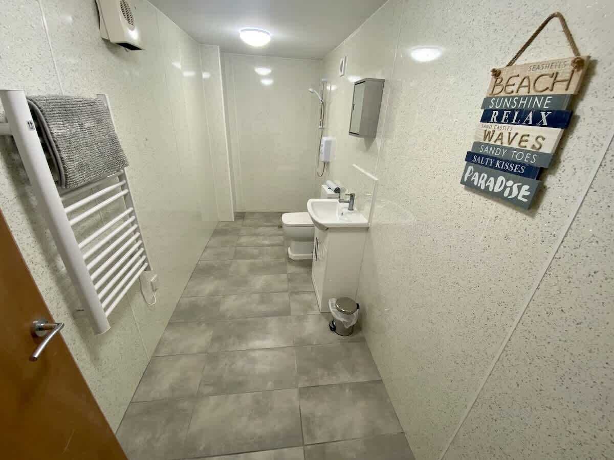 A narrow bathroom with a toilet, sink, and towel rack. A sign with words like 'beach' and 'sunshine' is visible.
