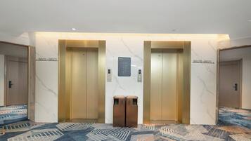 A hotel lobby with two elevators, a patterned carpet, and a wall-mounted information board.