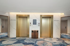 Vestíbulo de hotel con dos ascensores, alfombra estampada y un panel informativo en la pared.