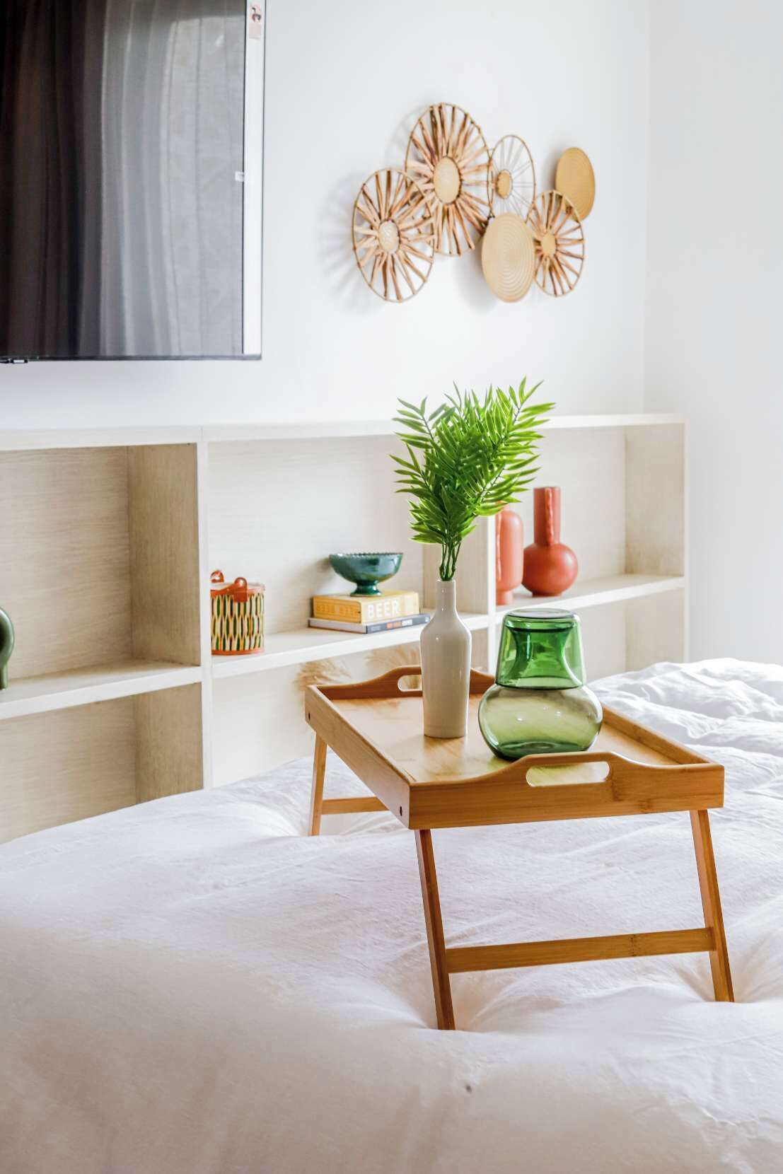 A bedroom with a bed, a wooden bedside table with a vase and a green glass, and a wall with decorative wooden items.