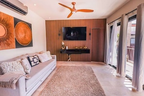 Ein modernes Wohnzimmer mit Sofa, Fernseher, Deckenventilator und einem großen Fenster mit Vorhängen.