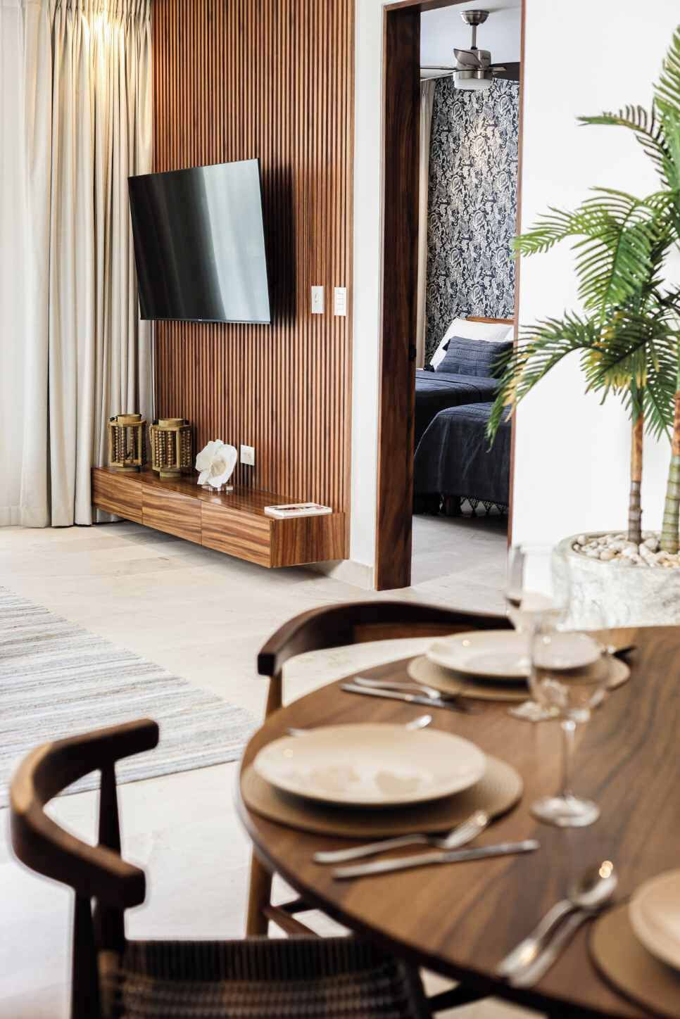 Ein modernes Wohnzimmer mit einem an einer Holzwand montierten Flachbildfernseher, einem Essbereich mit Tisch für zwei Personen und einem durch eine offene Tür sichtbaren Schlafzimmer.