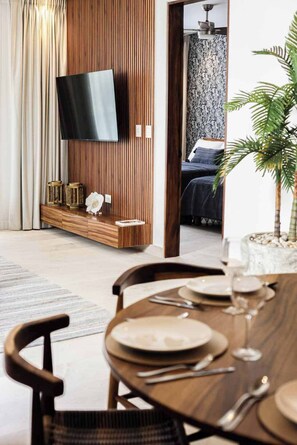 Een moderne woonkamer met een flatscreen televisie aan een houten wand, een eethoek met een tafel gedekt voor twee personen, en een slaapkamer zichtbaar door een open deur.