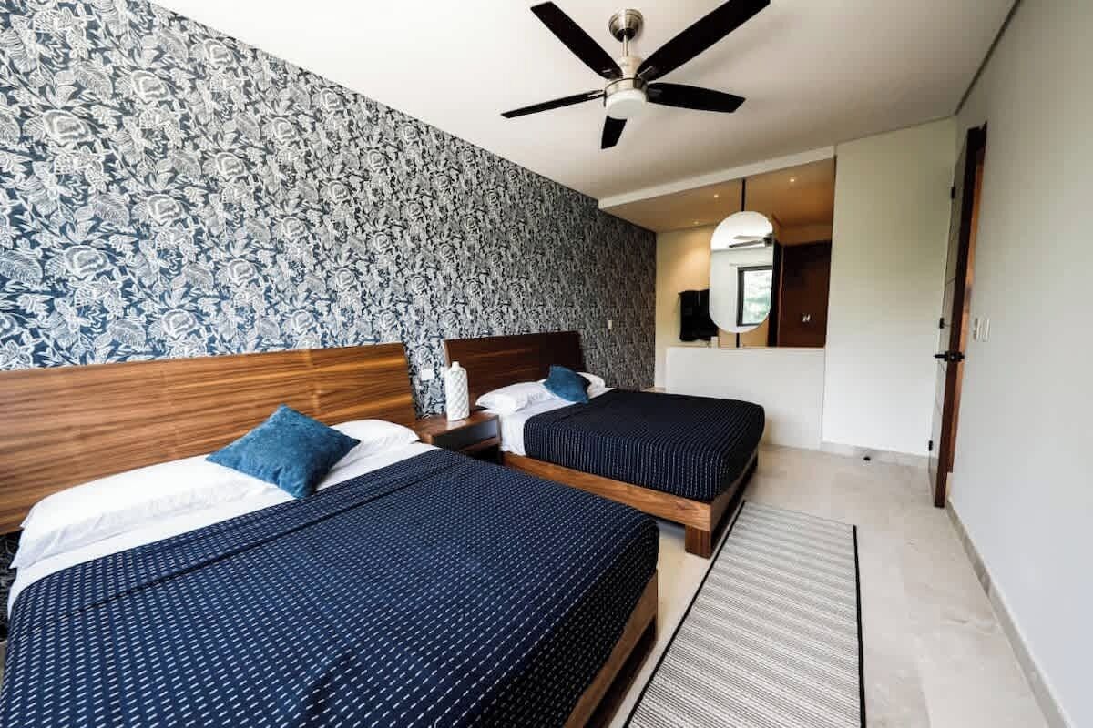 A hotel room with two beds, a ceiling fan, a patterned wallpaper, a mirror, and a wooden headboard.