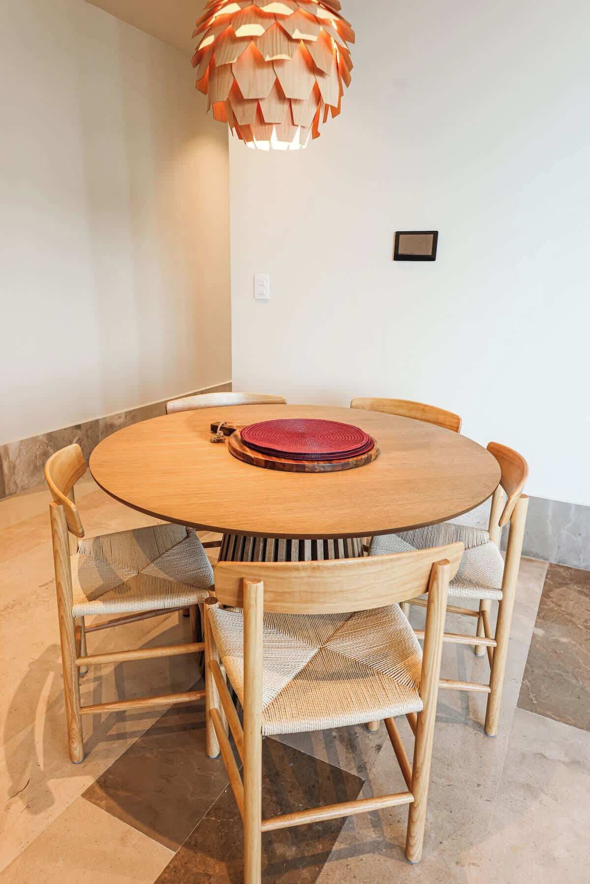 A round wooden dining table with woven chairs, a red placemat, and a unique paper cone chandelier.