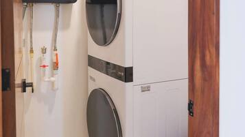 A laundry room with two stacked washing machines, a water heater, and a trash bin.