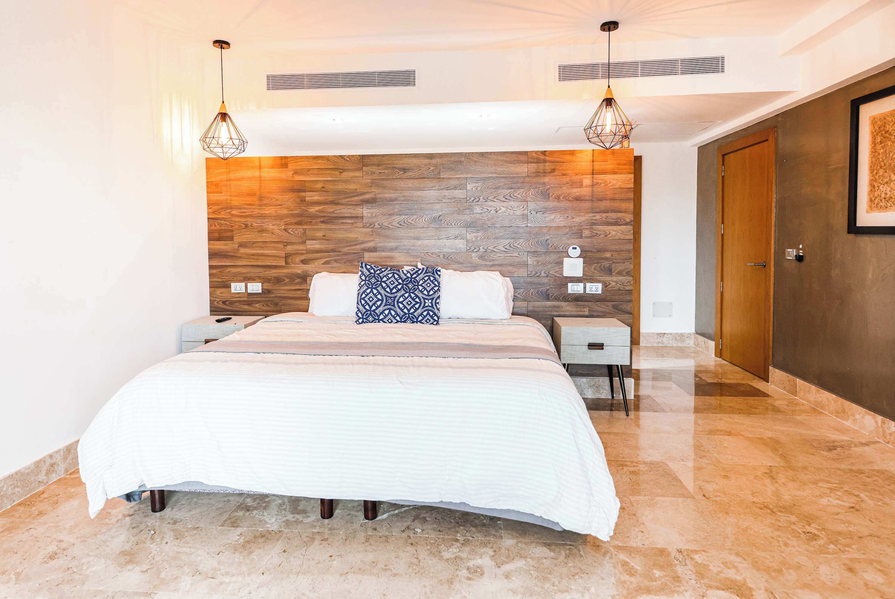 Quarto moderno com cabeceira de madeira, uma cama grande com colcha branca, duas mesas de cabeceira e um criado-mudo.
