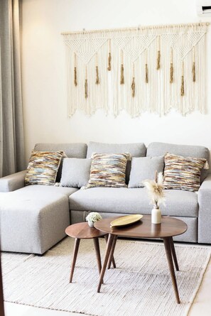 Una sala moderna con un sofá gris, cojines estampados, un colgador de macramé y una mesita de madera con un jarrón y un plato.