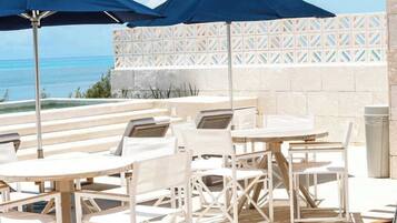 An outdoor seating area with tables and chairs, two umbrellas, and a view of the ocean.