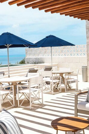 An outdoor seating area with tables and chairs, two umbrellas, and a view of the ocean. - Apt Jaguarundi: Stylish Retreat with Shared Pool (Puerto Morelos)