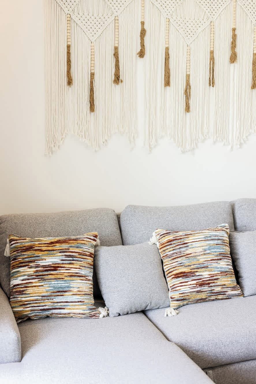 A grey sofa with two striped pillows and a macrame wall hanging above.
