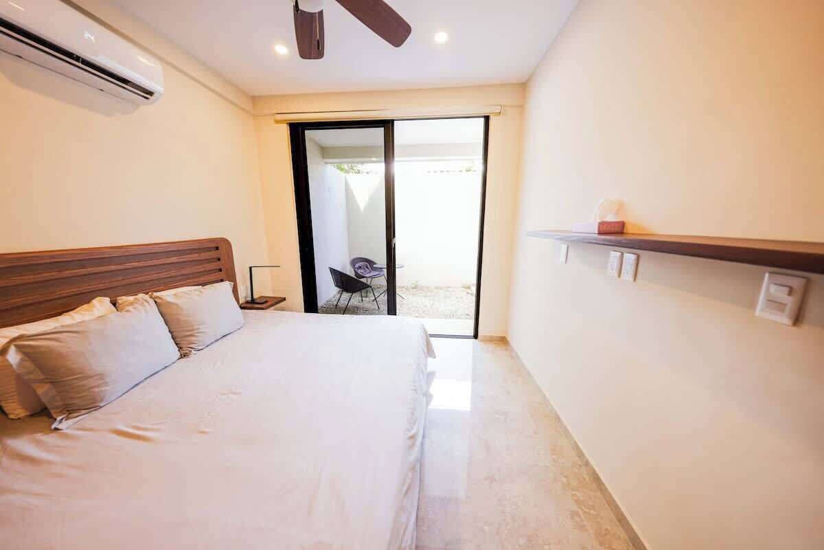 A hotel room with a bed, a ceiling fan, an air conditioning unit, a sliding glass door leading to an outdoor area, and a small table with a chair.