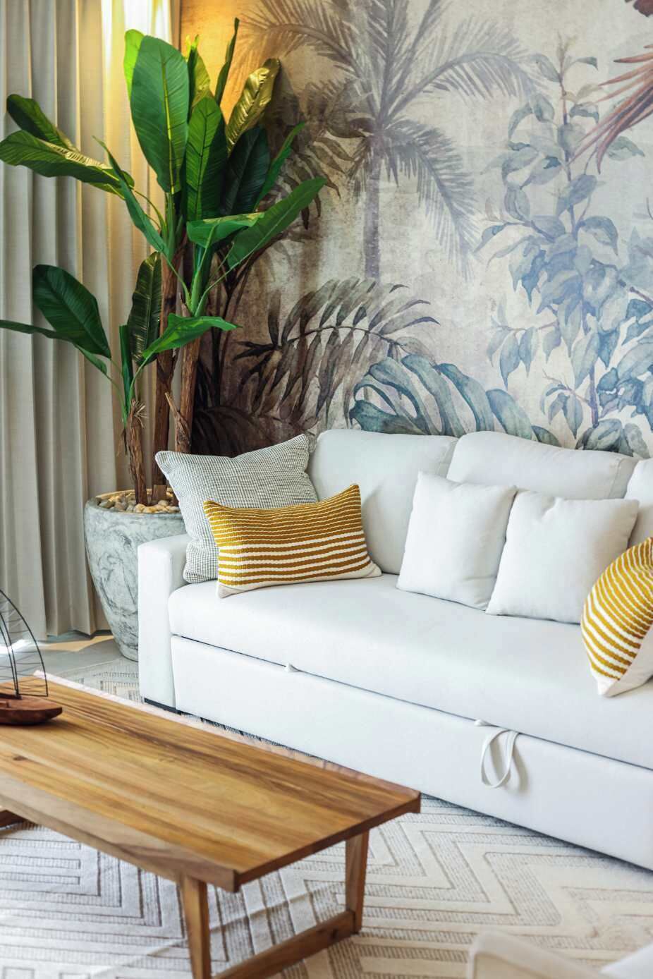 En hvid sofa med stribede puder, en stor grøn plante og en væg med et tropisk muralmaleri.