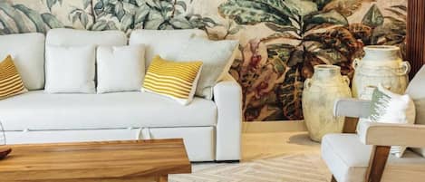 Un salon avec un canapé blanc, des coussins à rayures, une table basse en bois et une grande plante, le tout sur fond d’une fresque aux motifs tropicaux.